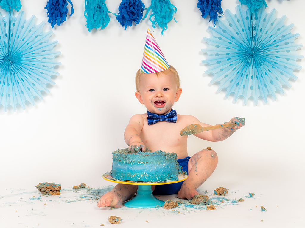 baby boy wearing party pat smashing blue cake with wooden spoon taken by cake smash photographer in Braintree